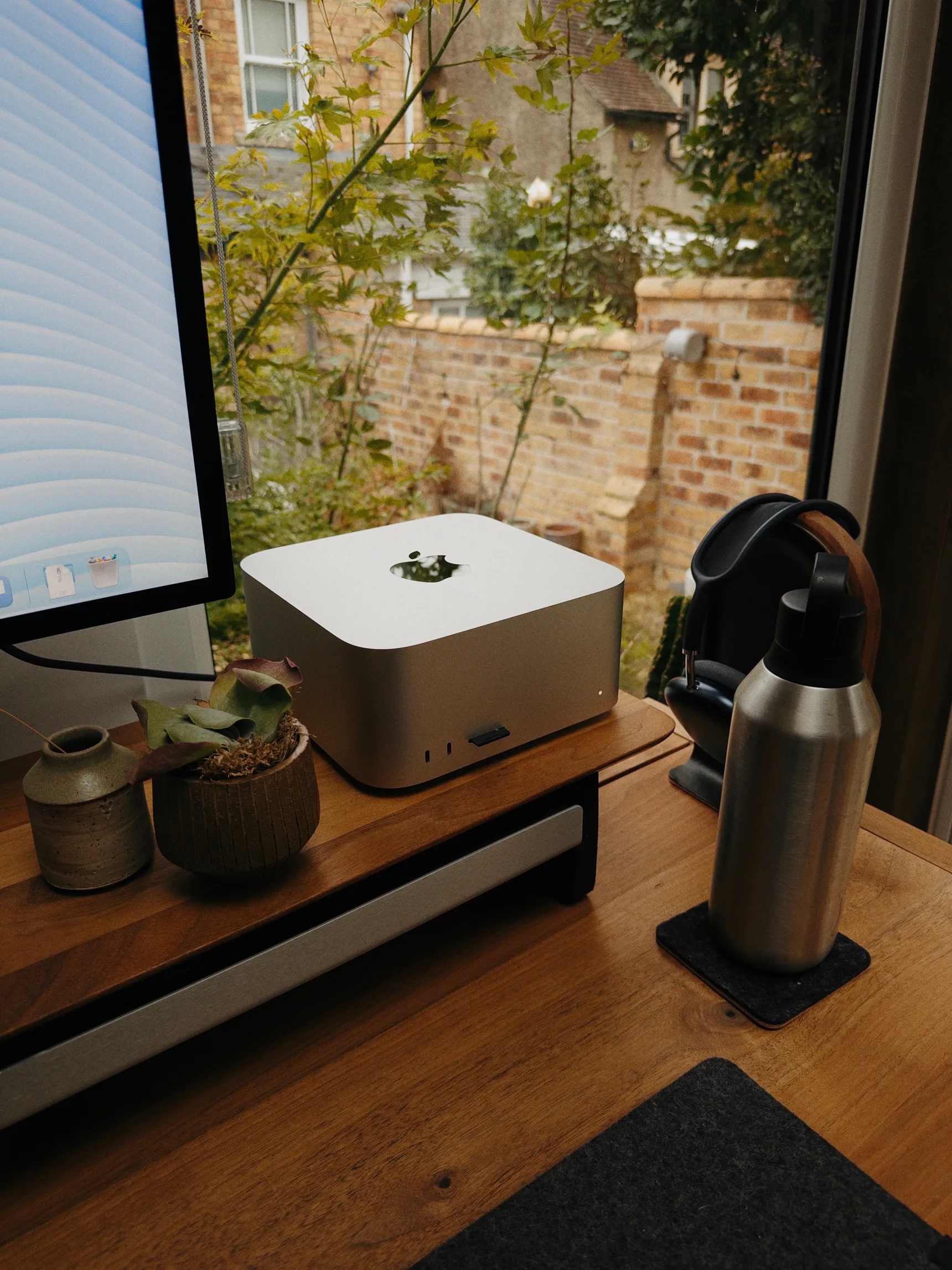 Mac Studio on a wooden desk by a window, beside a monitor and small potted plant. Headphones and a metal bottle sit to the right.